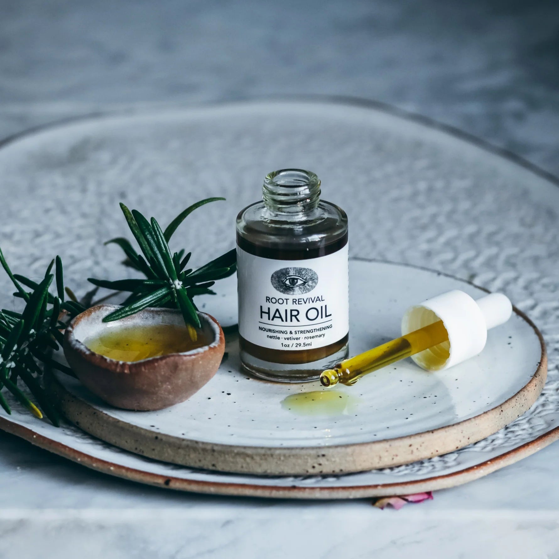 Bottle of hair oil with a dropper on a ceramic plate with rosemary and a small bowl of oil.