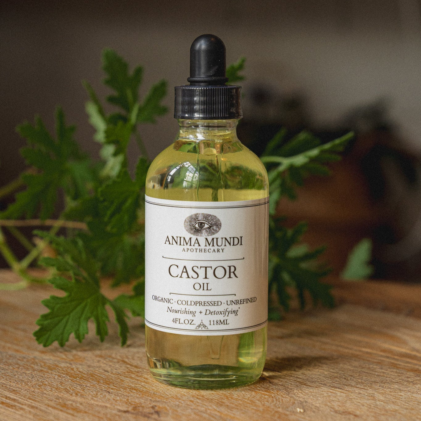 Bottle of Anima Mundi Castor Oil with a dropper on a wooden surface with green leaves.