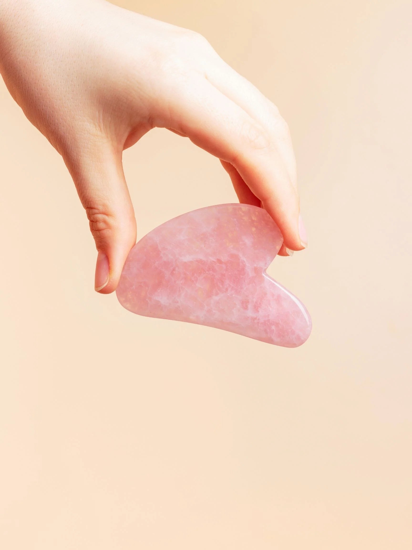 Hand holding a pink gua sha tool against a beige background