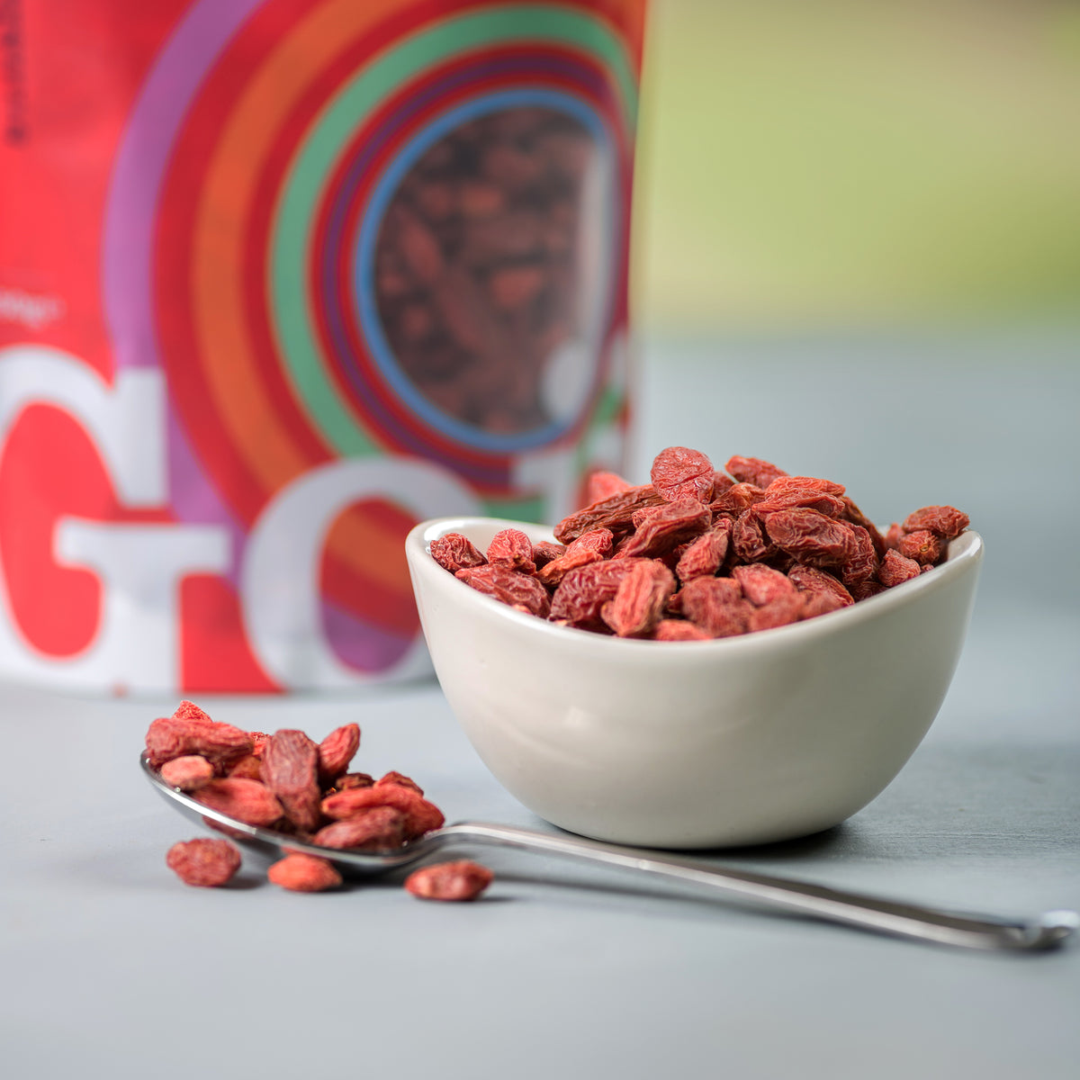 Bowl of goji berries with a spoonful on a gray surface, with a colorful box in the background.