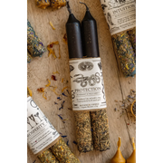 Two black candles with labels on a wooden surface with herbs and other items.