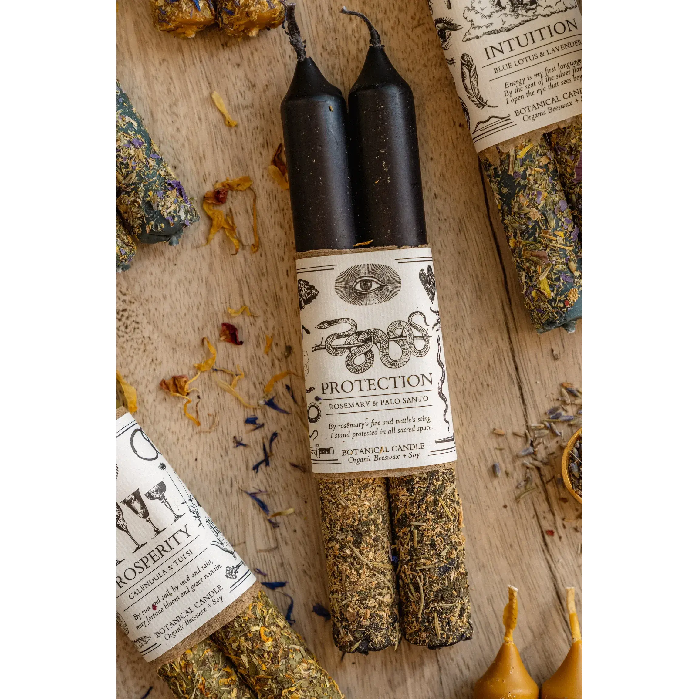 Two black candles with labels on a wooden surface with herbs and other items.
