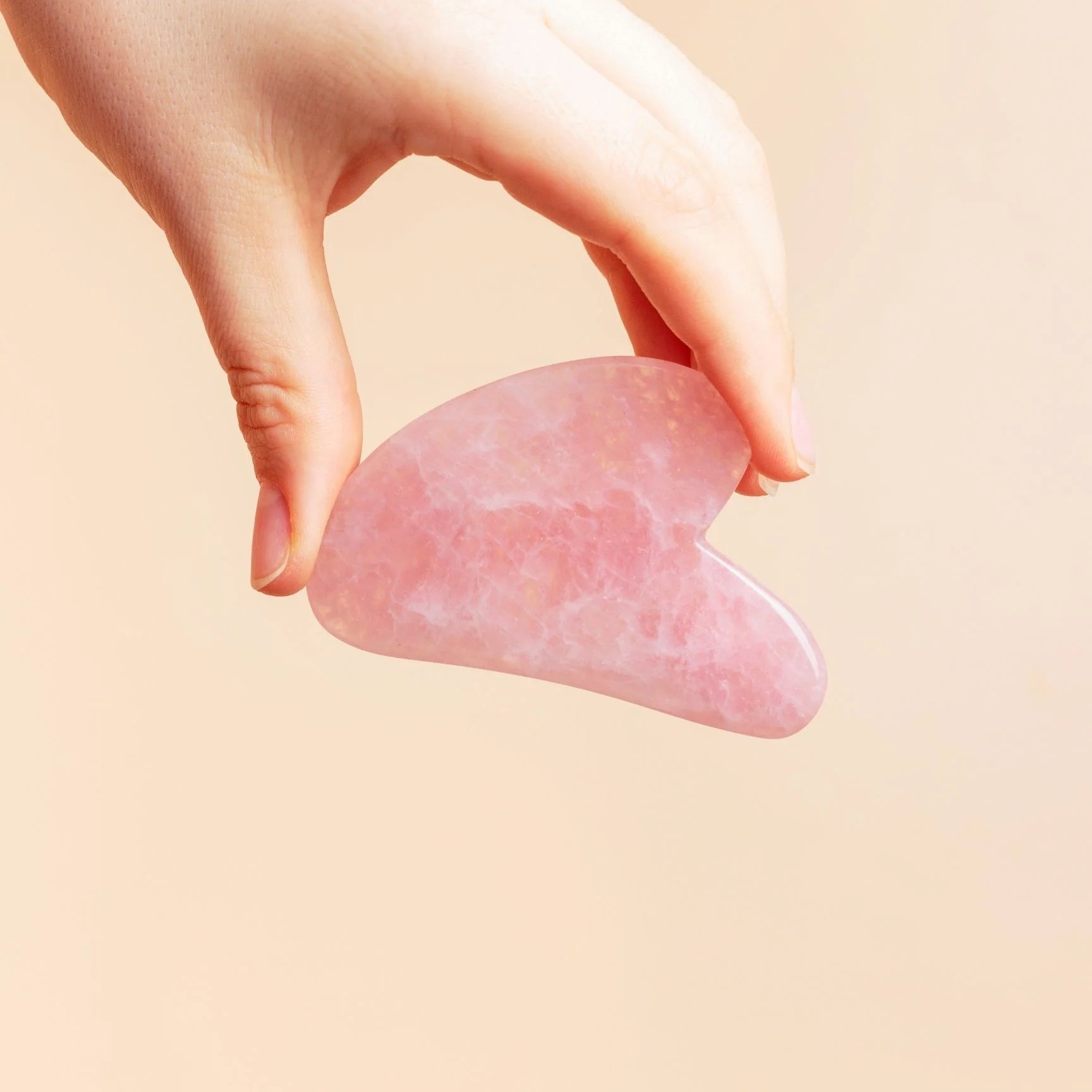 Hand holding a pink gua sha tool against a beige background