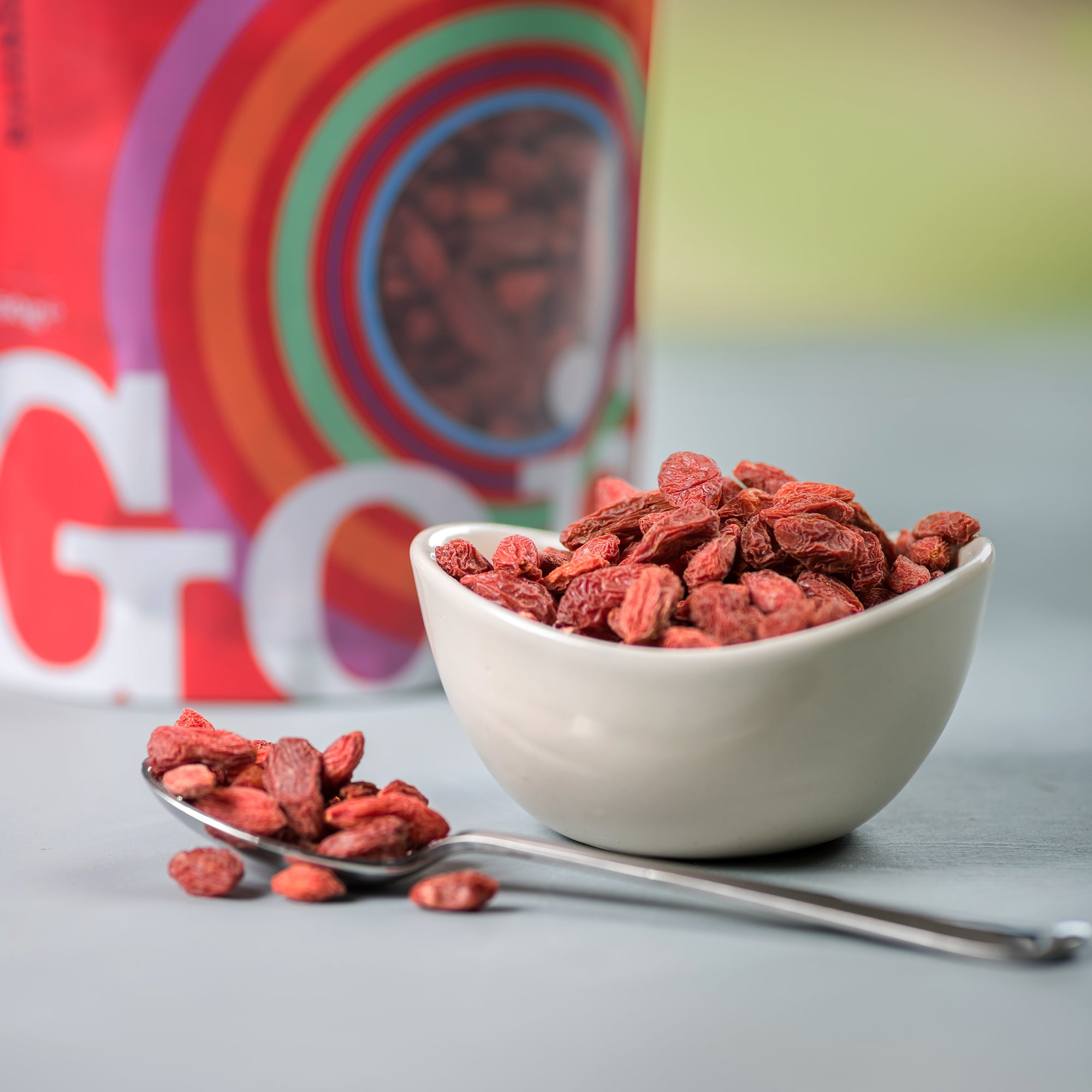 Bowl of goji berries with a spoonful on a gray surface, with a colorful box in the background.