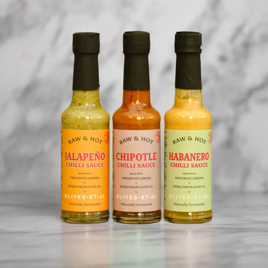 Three bottles of Raw & Hot chili sauces on a reflective surface with a marble background