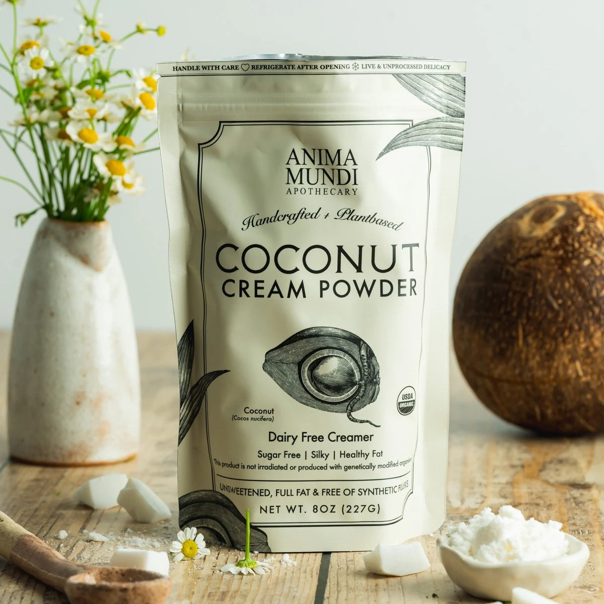 Anima Mundi Coconut Cream Powder pouch beside a whole coconut, daisies and coconut pieces