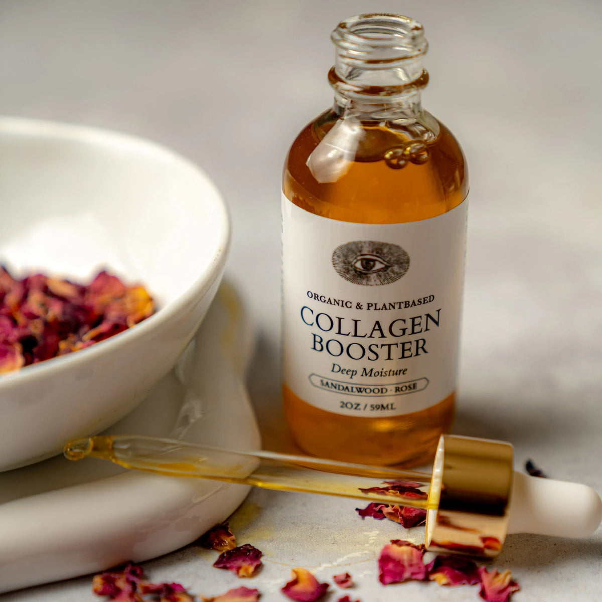 Anima Mundi Collagen Booster Face Oil bottle with its dropper, dried rose petals and a stone bowl