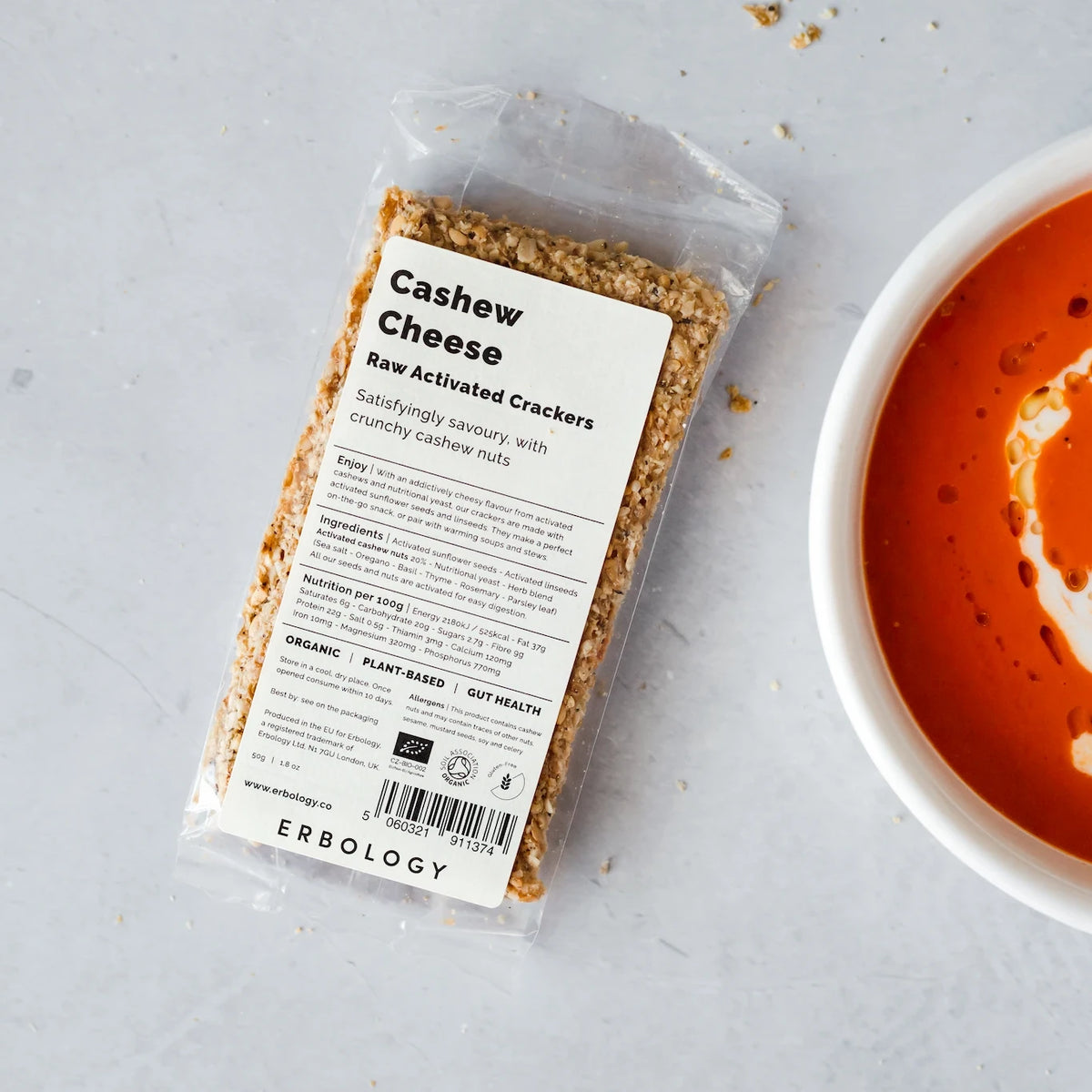 Pack of Erbology Cashew Cheese Raw Activated Crackers on a grey stone surface beside a bowl of tomato soup
