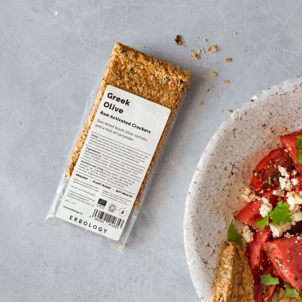 Pack of Erbology Greek Olive Raw Activated Crackers on a grey stone surface beside a bowl of tomato salad