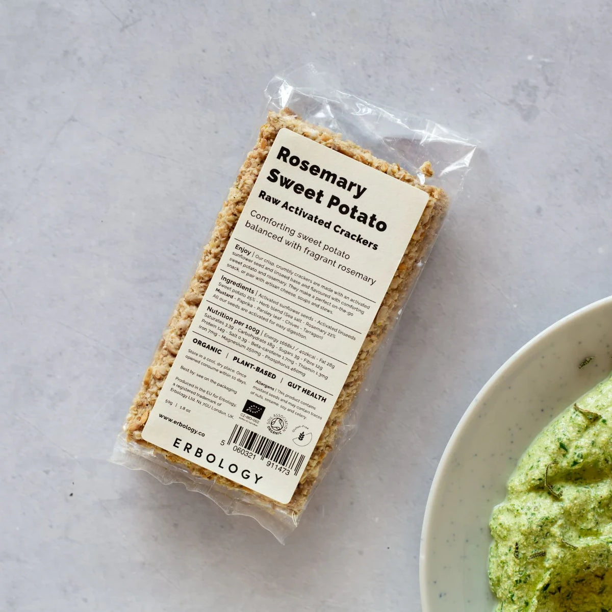 Pack of Erbology Rosemary Sweet Potato Raw Activated Crackers on a grey stone surface beside a bowl of green herb dip