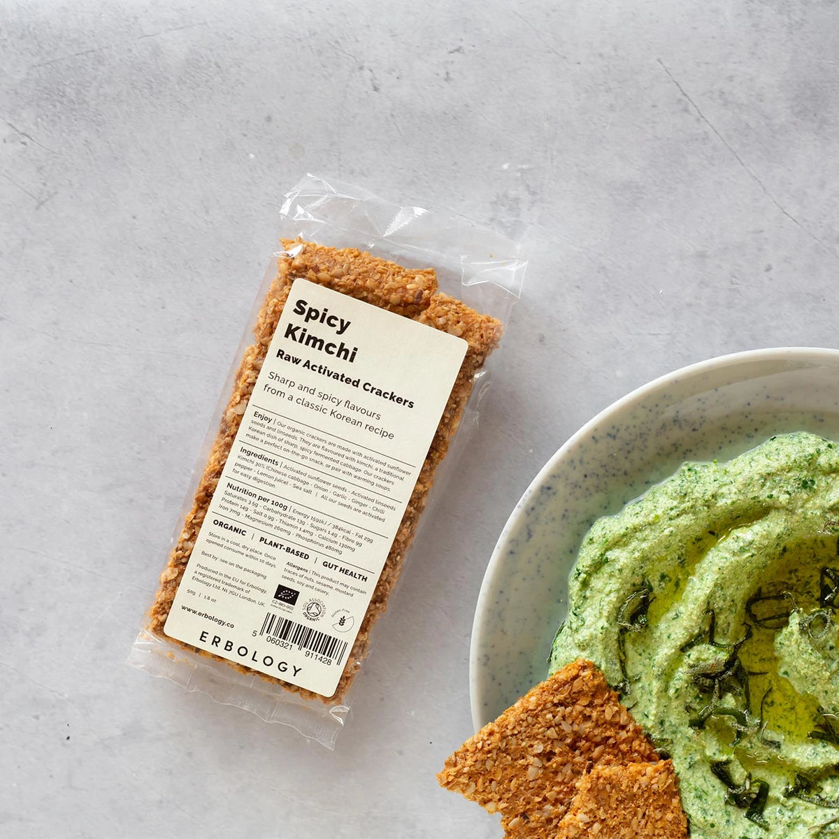 Pack of Erbology Spicy Kimchi Raw Activated Crackers on a grey stone surface beside a bowl of green dip with crackers