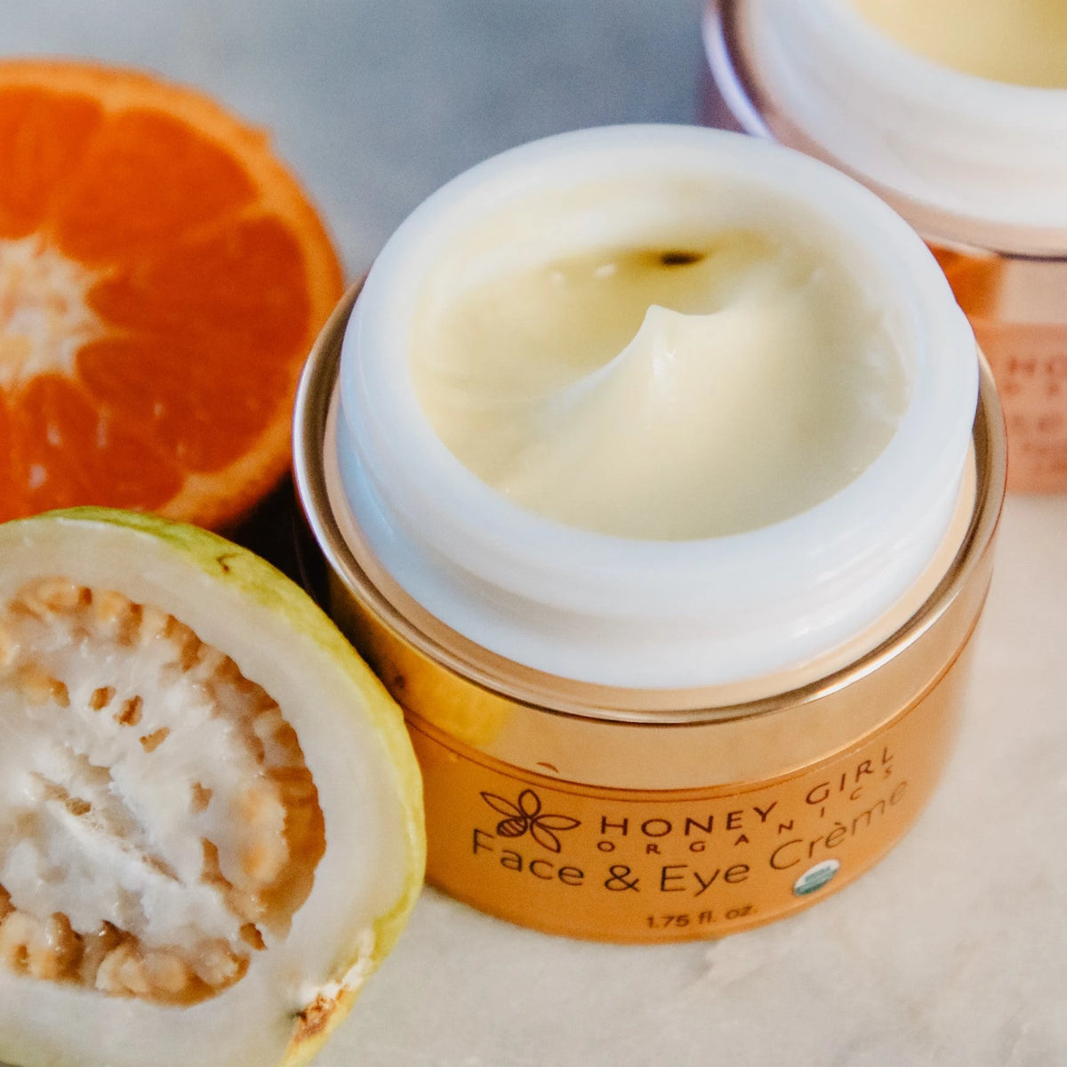 Open jar of Honey Girl Organics Face & Eye Crème with orange and guava halves