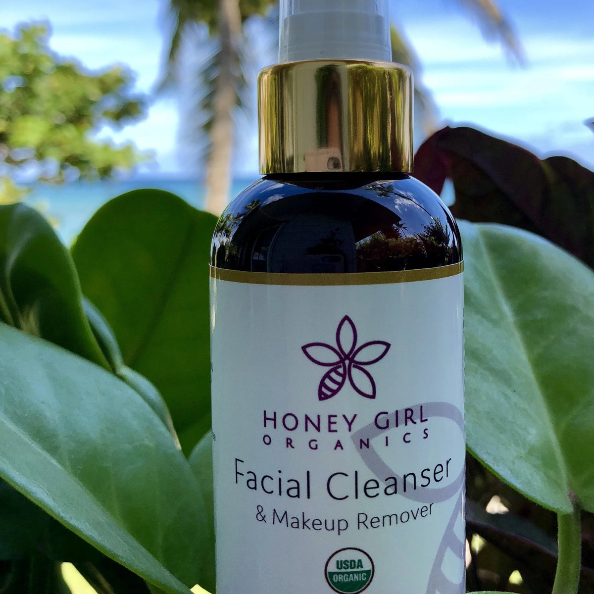 Bottle of Honey Girl Organics Facial Cleanser & Makeup Remover among green leaves and palm trees