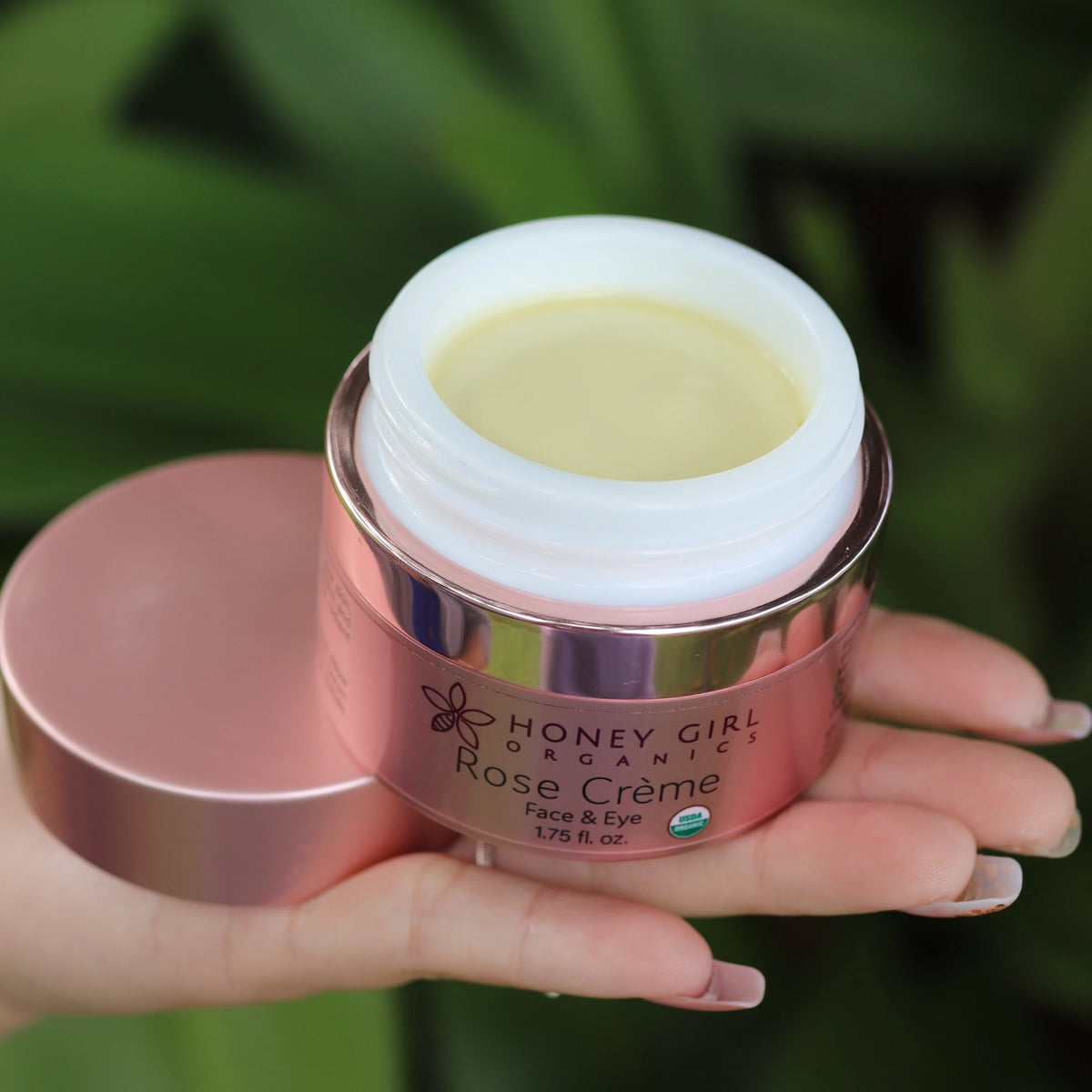 Open jar of Honey Girl Organics Rose Crème held in a hand against green leaves
