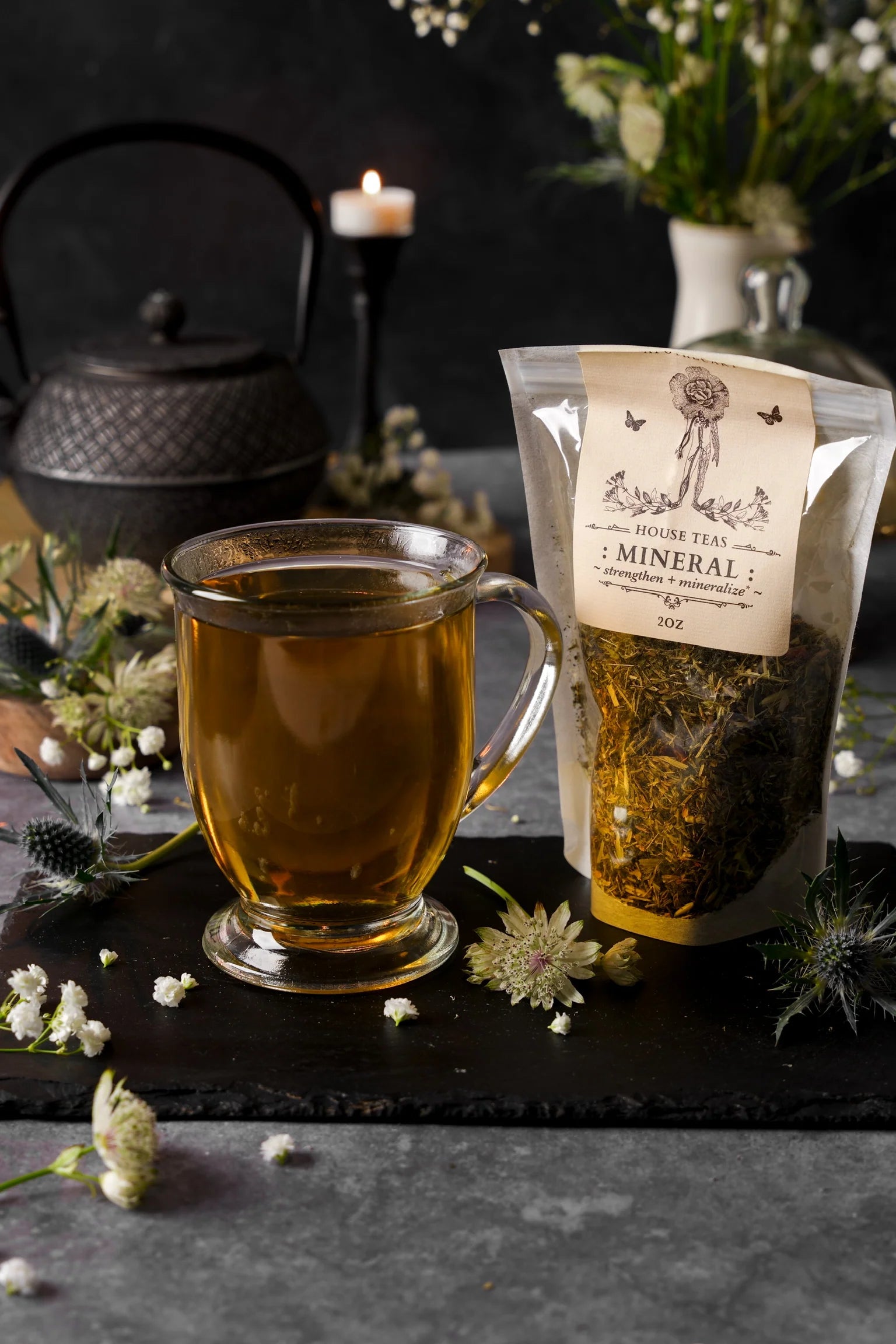 Tea cup with brewed tea next to a package of 'Mineral' tea on a dark surface with flowers and a candle in the background.