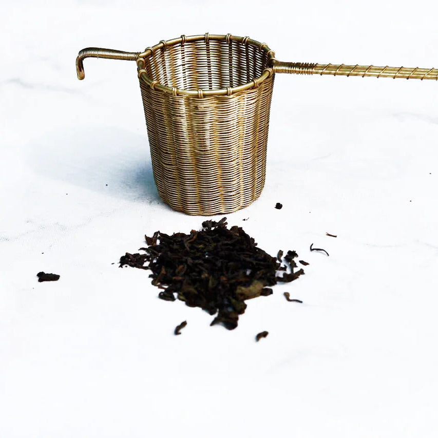 Woven brass tea strainer with loose tea leaves on a white background